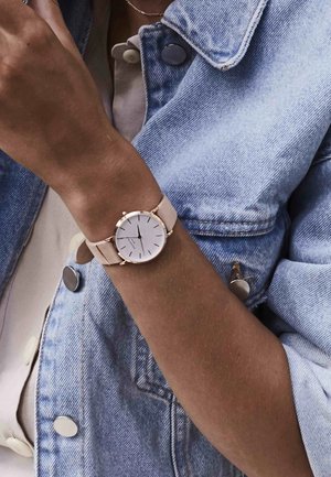 Kružni sat s bijelim brojčanikom, zlatnim detaljima i svijetlosmeđim kožnim remenom, nošen na zapešću s vidljivom traper jaknom.