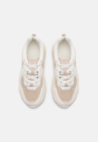 Bílé a béžové tenisky s prodyšným a hladkým materiálem, zaobleným tvarem, silnou podrážkou, bílými tkaničkami a logem uvnitř.