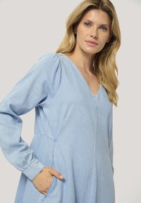 Světle modré šaty z chambray s dlouhými rukávy, výstřihem do V a nařasením na ramenou. Obsahují boční kapsy a mají volný střih.