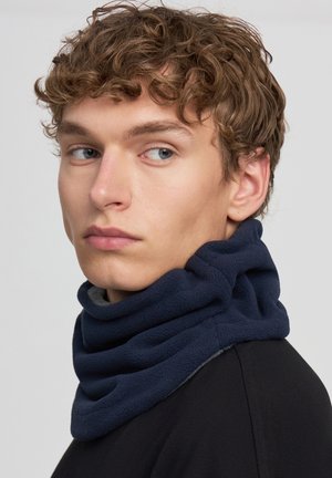 Giovane uomo con i capelli ricci che indossa un scaldacollo in pile blu navy, mentre guarda a sinistra, su uno sfondo chiaro e semplice.