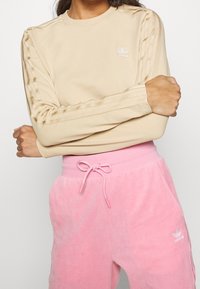 Beige långärmad topp med subtil logotyp och tre vertikala vita ränder på ärmarna, i kombination med mjuka rosa mjukisbyxor med dragsko i midjan.