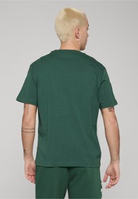 Ciemnozielona koszulka z krótkim rękawem wykonana z bawełny, z okrągłym dekoltem i luźnym krojem. Tył ma prosty design bez grafik.