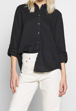 Person iført en sort langærmet skjorte med knapper og op rullede ærmer samt højtaljede off-white bukser, stående mod en neutral baggrund.