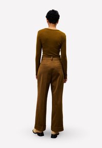 Persoon met kort krullend haar, gekleed in een aansluitend olijfgroene lange mouwen top en bruine high-waisted wijde broek, staande met de rug naar de camera gericht.