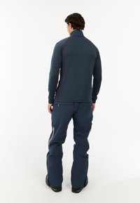 Marineblaues Langarmoberteil mit hohem Kragen, versehen mit roten Seitenelementen, kombiniert mit marineblauen Hosen und dunklen Schuhen, von hinten dargestellt.