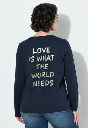 Donna con capelli corti e ricci che indossa una maglia a maniche lunghe blu navy con "LOVE IS WHAT THE WORLD NEEDS" stampato sul retro in lettere chiare.