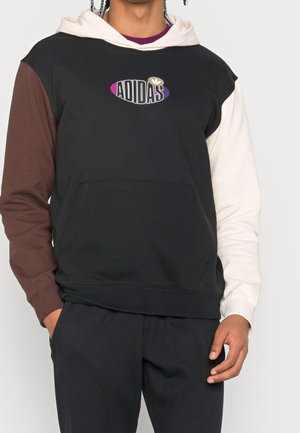 Hombre vistiendo una sudadera con capucha negra con una manga marrón, una manga blanca, capucha beige y logo multicolor de Adidas en el pecho.