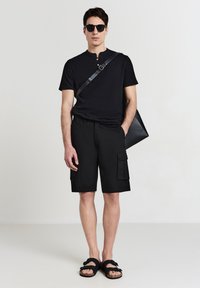 Uomo che indossa occhiali da sole neri, una maglietta henley nera a maniche corte, pantaloni cargo neri, sandali neri e porta una borsa a tracolla in pelle nera.