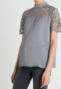 Blusa gris con cuello alto y mangas cortas, con detalle de encaje en los hombros y la parte superior, complementada con una suave tela de satén.