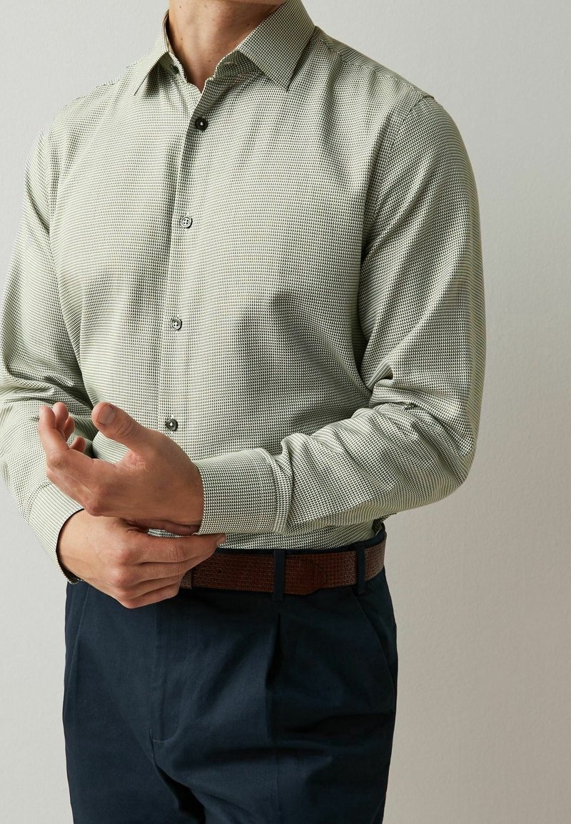 Homme ajustant le poignet d'une chemise à manches longues à motif clair rentrée dans un pantalon sombre avec une ceinture marron sur fond uni.