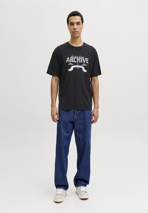 Man standing, wearing black graphic "Archive" T-shirt, loose blue jeans, and white sneakers, against a plain light gray background.