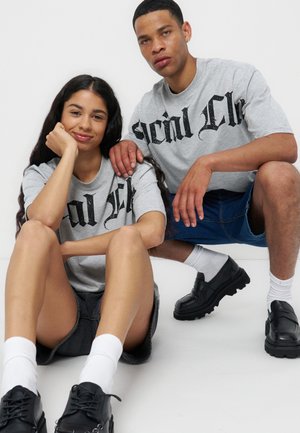 Ung kvinde sidder med bøjede ben og armen hvilende på knæet, ung mand sidder på hug ved siden af hende, begge iført grå T-shirts med grafik og sorte sko.