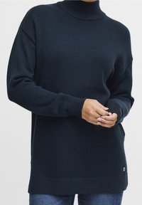 Granatowy, ciemny, dziany sweter z golfem o luźnym kroju, z prążkowanymi mankietami i brzegiem. Zawiera małe logo na dole po lewej stronie.