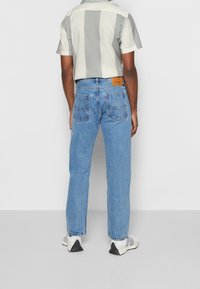 Homme portant un jean droit bleu clair, des baskets blanches et une chemise à manches courtes avec de larges rayures verticales grises et blanches, debout de dos.