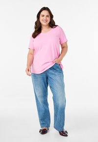 Ružičasta majica s V izrezom napravljena od mekog materijala, u kombinaciji s svijetlo plavim širokim trapericama i tamnim cipelama s dekorativnom kopčom.