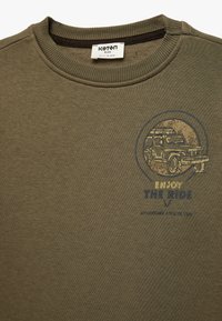 Olijfgroene sweater met ribbelachtige halslijn, voorzien van een graphic van een voertuig en de tekst "ENJOY THE RIDE" in blauw en geel. Zacht, gestructureerd stof.