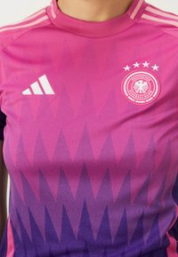 Koszulka reprezentacji Niemiec w gradientowych odcieniach różowego i fioletowego, z geometrycznym wzorem, logo Adidasa oraz herb drużyny z czterema gwiazdkami.