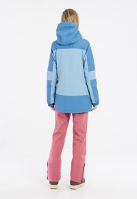 Blauwe en lichtblauwe waterdichte jas met een capuchon, elastische manchetten en gespleten naden, gecombineerd met roze broek en beige schoenen.