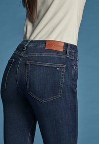 Person trägt dunkelblaue Tommy Hilfiger Jeans und ein hellbeiges Oberteil vor einfarbig blauem Hintergrund.