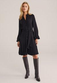 Donna che indossa un vestito lungo a maniche nere con bottoni e cintura in vita, e stivali alti fino al ginocchio di un marrone scuro, in piedi contro uno sfondo semplice.