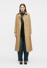 Γυναίκα που φοράει ένα μακρύ μπεζ καμπαρντίνα πάνω από γκρι πουλόβερ ζιβάγκο, παντελόνι με φαρδύ πόδι και καφέ σκούρα παπούτσια, στέκεται μπροστά σε ένα μονόχρωμο φόντο.