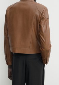 En person klädd i en brun läderjacka och svarta byxor, sedd bakifrån mot en enfärgad ljus bakgrund.