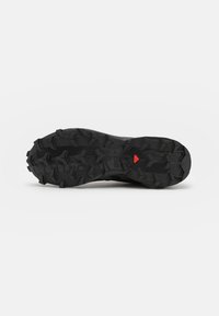 Semelle d'une chaussure de sport noire avec un motif de crampons profonds et robustes et un petit détail triangulaire rouge au centre.
