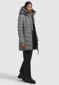 Woman wearing a long gray padded coat with a large hood, black pants, and black chunky boots, standing against a plain light background.