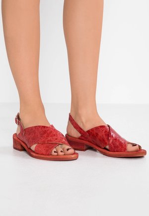 Pieds chaussés de sandales basses en cuir rouge avec des brides croisées et des orteils ouverts sur un sol blanc et un fond blanc.