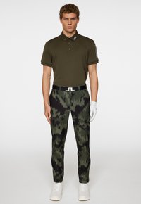 Olivgrön polo t-shirt, svarta mönstrade byxor med gröna detaljer, vit bälte, vita skor och en vit golfhandske.