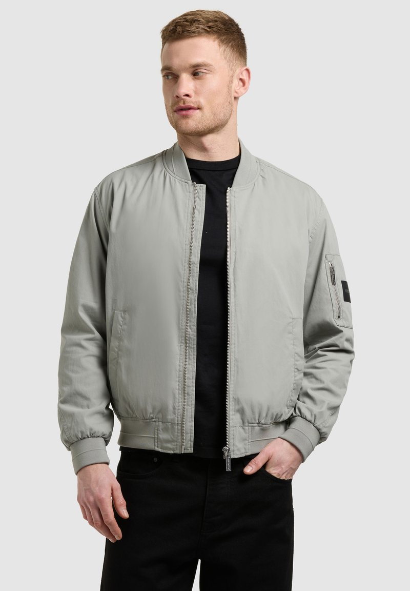 Homme portant une veste bomber gris clair par-dessus une chemise noire et un pantalon noir, regardant légèrement vers la gauche avec la main droite dans la poche.