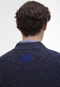 Marine gebreide trui met een textuurpatroon, voorzien van een onderscheidende blauwe patch bij de achterkant van de halslijn en een geribde kraag.