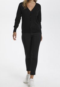 Svart cardigan med v-ringad halsringning, med knappar framtill. Tillsammans med svarta kroppsnära byxor och vita sneakers. Slät textur.