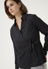 Camicia nera a portafoglio con pattina dei bottoni nascosta, maniche lunghe e dettaglio con laccio in vita, realizzata in tessuto leggero e liscio.
