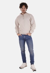 Felpa beige con zip in morbido tessuto, abbinata a jeans skinny blu e sneakers chiare, in piedi contro uno sfondo neutro.