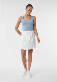 Hellblauer ärmelloser Bodysuit kombiniert mit einem weißen Wickelrock mit Bindegürtel. Das Modell trägt weiße Sneaker; glattes Material, lockere Passform.