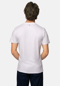 Mand med brunt hår iført en almindelig hvid t-shirt med korte ærmer og mørkeblå jeans, set fra ryggen på en hvid baggrund.