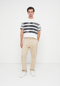 Λευκό κοντομάνικο μπλουζάκι με ναυτικές οριζόντιες ρίγες και το λογότυπο 'GUESS', συνδυασμένο με μπεζ παντελόνι και λευκά αθλητικά παπούτσια. Απλή, καθημερινή εμφάνιση.