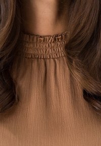 Blouse brun à texture avec un col froncé. Le tissu semble léger et légèrement froissé, mettant en valeur sa forme douce et fluide.