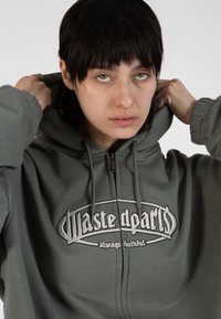 Pilkos zip-up tipo gobtuvu striukė su dideliu išsiuvinėtu "Waste parts" logotipu. Pasižymi gobtuvu su raišteliais ir įlenktais rankogaliais, pagaminta iš minkštos medžiagos.