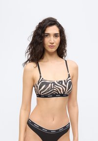 Γυναίκα με μακριά σγουρά μαλλιά που φοράει σουτιέν με μοτίβο ζέβρας και μαύρο σλιπ με το λογότυπο Hugo στις ελαστικές ταινίες, στέκεται μπροστά σε λευκό φόντο.