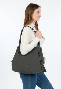 Donna con maglione bianco a coste e jeans blu che tiene una grande borsa tote grigio scuro con nappe sulla spalla, rivolta verso destra.