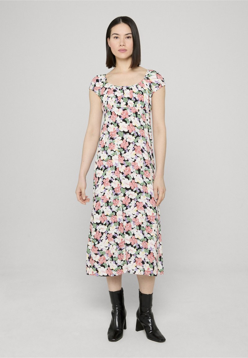 Donna in piedi che indossa un vestito floreale di media lunghezza con maniche corte e stivaletti neri alla caviglia, su uno sfondo semplice grigio chiaro.