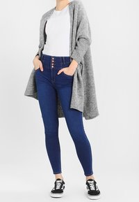 Harmaa neulottu auki oleva kardigan, valkoinen t-paita, siniset korkeavyötäröiset farkut, joissa on nappikoristeet, ja mustat tennarit valkoisilla pohjilla.