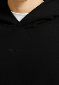 Svart hoodie i en bomullsblandning, med subtil upphöjd text "LOT: 3300/03" på framsidan. Mjuk textur med en avslappnad passform.