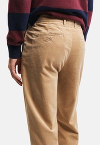 Beige Cord-Ripp-Hosen mit gerippter Textur, zwei Hintertaschen und einem taillierten Bund. Ein gestreifter maroon- und marineblauer Pullover ist teilweise sichtbar.