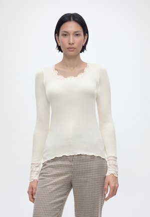 Donna con capelli corti e scuri che indossa una maglia crema a maniche lunghe con dettagli in pizzo e pantaloni a quadri, in piedi davanti a uno sfondo chiaro e uniforme.