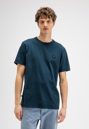 Junger Mann mit lockigem Haar, der ein dunkelblaues T-Shirt mit kurzen Ärmeln und helle blaue Jeans trägt, steht vor einem schlichten weißen Hintergrund.