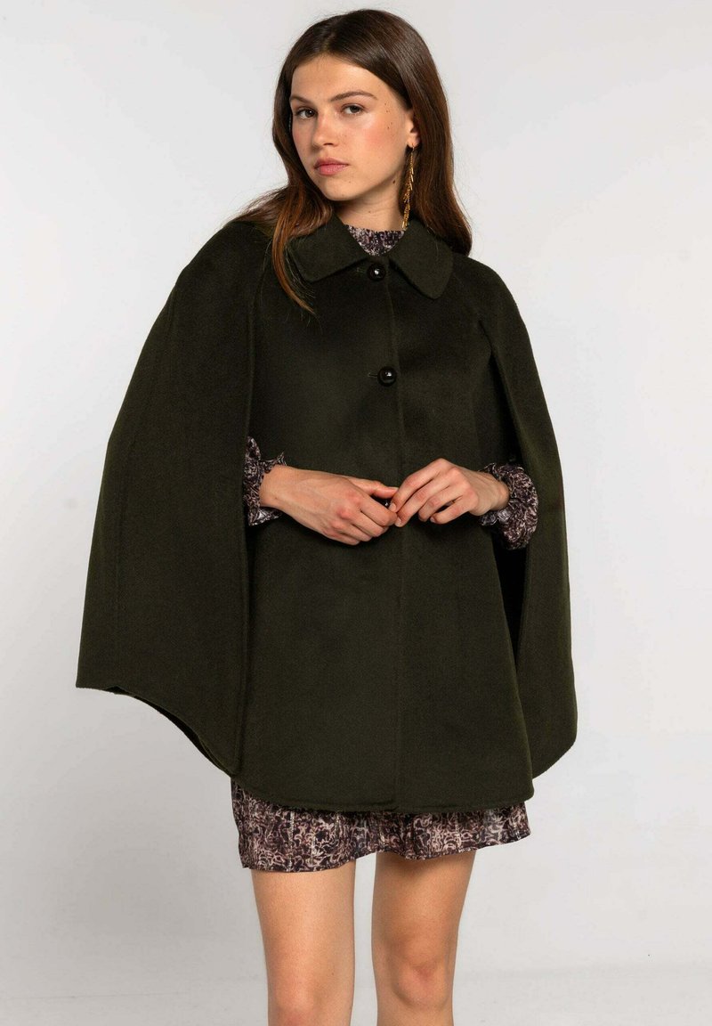 Cape vert foncé avec un col, dotée d'une fermeture à bouton et d'un design fluide. Associée à une robe à motif, le look global est superposé.