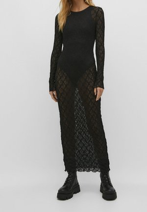 Vrouw die een lange mouwen zwarte kanten jurk draagt over zwarte onderkleding en grove zwarte veterlaarzen, staand tegen een effen achtergrond.